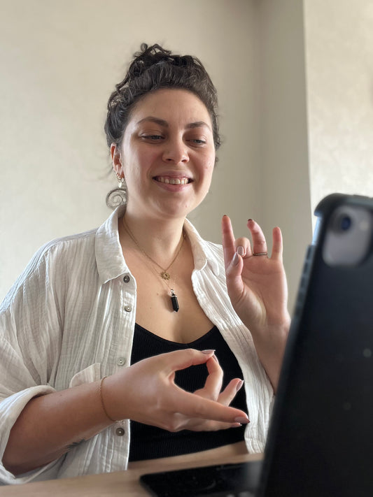 Femme souriante assise devant un ordinateur, effectuant un geste de méditation ou de transmission énergétique avec les mains. Elle porte une chemise blanche ouverte sur un haut noir, un pendentif en pierre naturelle et des boucles d’oreilles dorées. L’ambiance douce et naturelle reflète un moment de guidance, d’enseignement ou de connexion spirituelle à distance.